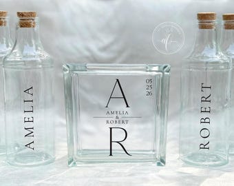 May include: Clear glass bottles with cork stoppers and personalized text. A glass block with the initials "A" and "R" and the date "05 25 26" is centered. The bottles are labeled "AMELIA" and "ROBERT".