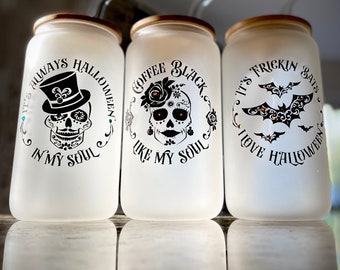 May include: Three frosted glass tumblers with gold lids and black and white Halloween-themed designs. The designs include a skull with a top hat, a skull with a flower, and a bat. The text on the tumblers reads "It's always Halloween in my soul", "Coffee black like my soul", and "It's frickin' bats I love Halloween".