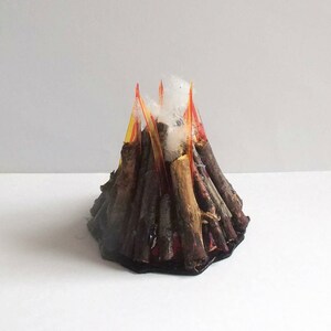 May include: A handmade volcano model with a dark brown base and sides, featuring red, orange, and yellow flames at the top. White fluffy material simulates smoke. The volcano is set against a plain white background.