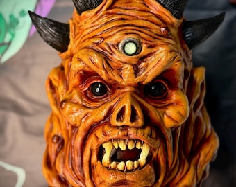 May include: A detailed orange and brown demon mask with black horns, sharp teeth, and a third eye. The mask has a textured surface and is designed with intricate details, perfect for Halloween or cosplay.