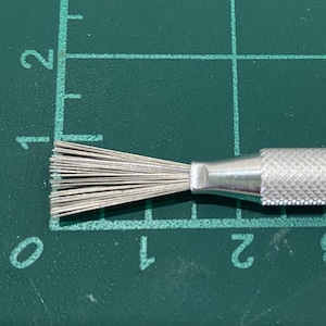 May include: A silver cleaning tool with a brush on one end and a textured handle. The brush has a fan shape with many fine bristles.