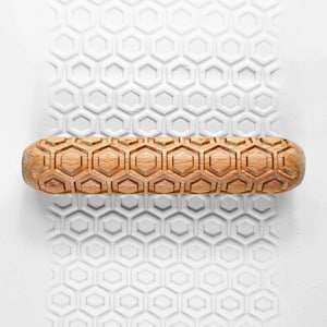 May include: A wooden rolling pin with a honeycomb pattern is shown on a white surface with a matching embossed design. The rolling pin is light brown and cylindrical, with a repeating geometric pattern carved into its surface. The white surface has a similar pattern.