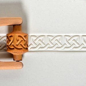 May include: A light brown wooden massage roller with a carved Celtic knot pattern. The roller has two wooden handles. The background is a white surface with a repeating Celtic knot design.
