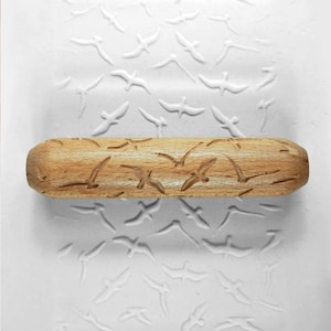 May include: A wooden rolling pin with a carved design of flying birds. The rolling pin is on a white surface with an embossed pattern of flying birds.