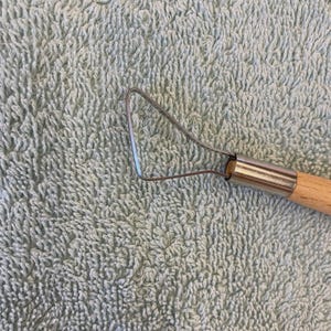 May include: A pottery ribbon tool featuring a triangular-shaped wire loop made of thin metal, securely attached to a smooth, light-coloured wooden handle with a silver-toned metal ferrule connecting the two components for sculpting clay.