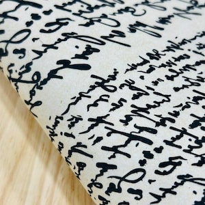 May include: A piece of off-white fabric featuring a repeating pattern of black handwritten cursive script. The text consists of stylised, illegible calligraphy that flows across the surface in dense, overlapping lines, creating a dark contrast.