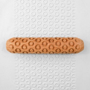 May include: A wooden rolling pin with a circular pattern embossed on the surface. The rolling pin is light brown in colour and is lying on a white surface with a repeating pattern of circles.