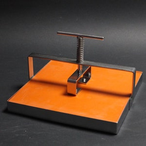 May include: A metal press with a square orange base and a metal handle with a spring mechanism. The press is used to flatten objects.
