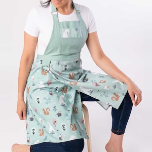 May include: A green apron with a white pocket and a woodland animal print featuring bears, squirrels, and owls. The apron has adjustable straps and a D-ring closure.