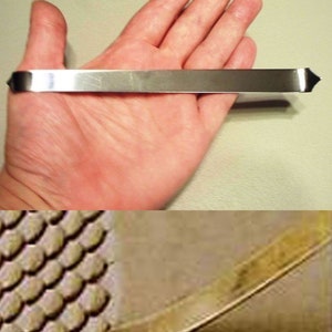 May include: A silver metal tool with a rounded end and a flat, rectangular shape. The tool is approximately 15 cm long and is held in a person's hand.