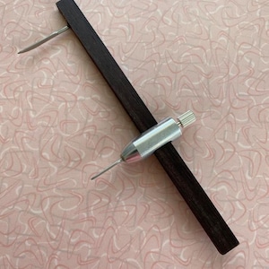 May include: A wooden and metal circle drawing tool with a pointed tip and a knob for adjusting the circle size. The tool is on a pink and white patterned surface.