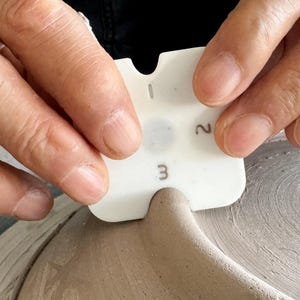 May include: A white pottery rib tool, marked with the numbers 1, 2, and 3, is used to shape wet clay. The tool is held by two hands. The clay is a light brown colour. The image shows a pottery process.