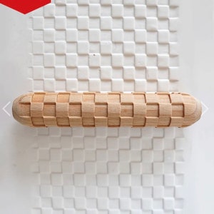 May include: A wooden rolling pin with a square patterned design. The rolling pin is made of light brown wood and has a smooth, rounded handle.
