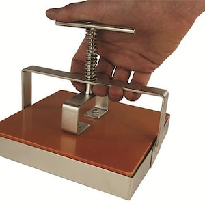 May include: A stainless steel press with a brown rubber pad and a spring-loaded handle. The press is used to flatten objects.
