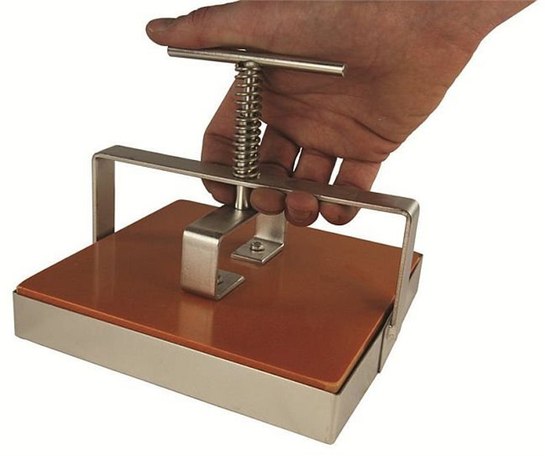May include: A stainless steel press with a brown rubber pad and a spring-loaded handle. The press is used to flatten objects.