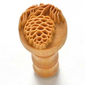 May include: A wooden stamp featuring a detailed design of a pine cone and leaves. The stamp is light brown and has a round top with a cylindrical handle.