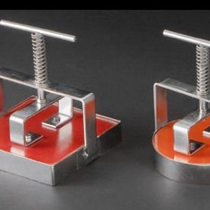 May include: Two stainless steel burger press tools with red non-stick surfaces. The larger press has a square surface and the smaller press has a round surface.