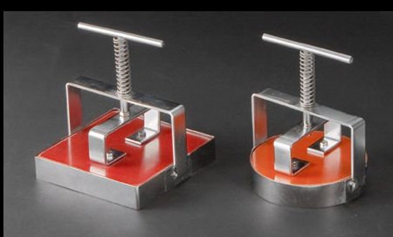 May include: Two stainless steel burger press tools with red non-stick surfaces. The larger press has a square surface and the smaller press has a round surface.
