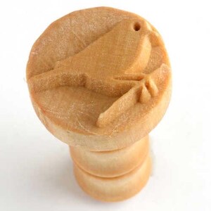 May include: A wooden stamp featuring a bird motif. The stamp has a detailed carving of a bird on a branch. The stamp is light brown in colour and has a rounded top with a handle made of stacked, rounded pieces.