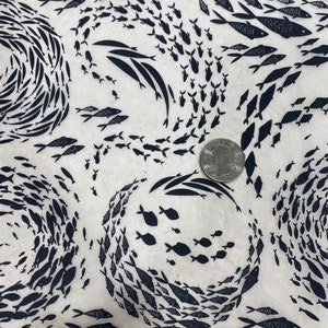 May include: Off-white fabric featuring a repeating pattern of black fish in swirling formations. The design includes various sizes of fish, creating a dynamic aquatic theme. A coin is included for scale.