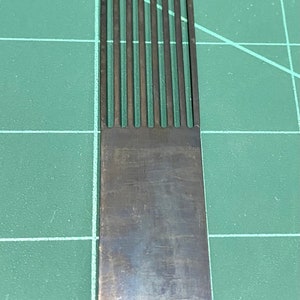 May include: A close-up of a metal comb with a row of thin, evenly spaced teeth. The comb is silver in colour and has a smooth, polished finish.