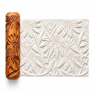 May include: A cylindrical wooden rolling pin with a carved floral pattern featuring coneflowers. Next to the rolling pin is a rectangular slab of white clay that has been impressed with the same floral design, showing the raised texture.