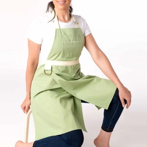 May include: A light green apron with a white adjustable strap and a single pocket. The apron is tied at the waist with a white strap.