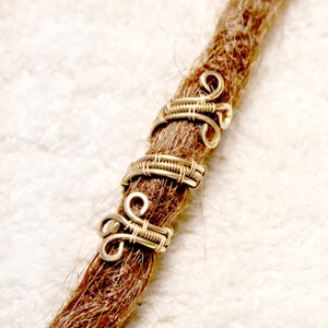 Pode incluir: Três contas de dreadlocks de fio dourado com um design decorativo em espiral. As contas estão em um dreadlock marrom.