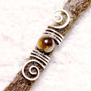 Könnte beinhalten: Ein silberfarbenes Haaraccessoire aus Metall mit einer braunen Edelsteinperle. Das Accessoire ist um einen braunen Dreadlock gewickelt. Das Design umfasst Spiralakzente und Drahtwicklungen, wodurch ein einzigartiger Haarschmuck entsteht.