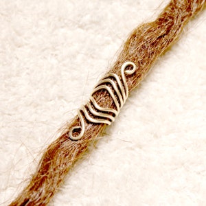 Op de afbeelding: Zilveren dreadlock-kraal met een zwierig, ingewikkeld ontwerp. De kraal is gemaakt van draad en is om een ​​bruine dreadlock gewikkeld.