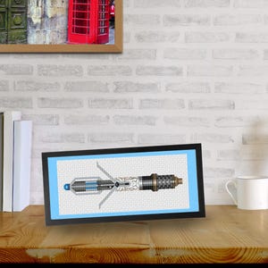 Peut inclure: Une œuvre d'art encadrée en point de croix représentant un tournevis sonique. Le design incorpore des nuances de bleu, d'argent et de noir, sur un fond bleu clair. L'œuvre est exposée sur une surface en bois.