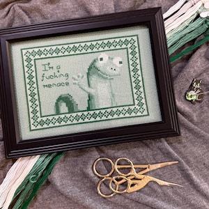 May include: Framed cross-stitch art featuring a green frog design and the text "I'm a fucking menace." The artwork is in a black frame. Gold scissors and green embroidery thread are also visible.