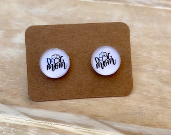 Puede incluir: Un par de pendientes de botón blancos con el texto "Dog Mom" y un diseño de huella de pata en negro.