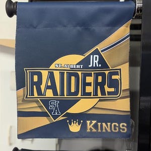 Puede incluir: Una pancarta azul marino con detalles dorados y blancos. La pancarta presenta el texto "ST. ALBERT RAIDERS JR. KINGS" en una fuente audaz y estilizada. También hay un gráfico de corona.