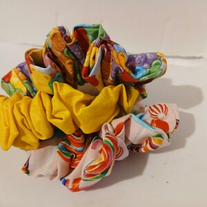 May include: A set of three fabric hair scrunchies stacked together. The top scrunchie features a colorful candy print, the middle one is a solid bright yellow with a subtle shimmer, and the bottom one has a light pink base with a candy pattern.