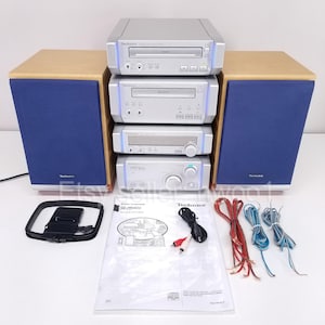 May include: A silver Technics stereo system with two speakers, a CD player, and an amplifier. The speakers have blue fabric fronts and wooden frames. Includes a manual, cables, and an antenna. The system is on a white surface.