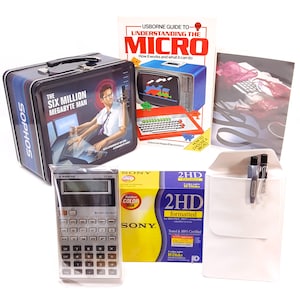 Può includere: Una vintage lunchbox in metallo con un design nero e blu che presenta il testo "The Six Million Megabyte Man" e un'immagine di un uomo dei cartoni animati che usa un computer. La lunchbox è accanto a un libro intitolato "Understanding the Micro" e una calcolatrice. Ci sono anche due penne, una busta bianca e una scatola di floppy disk formattati 2HD.