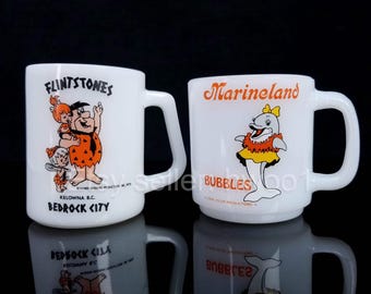 May include: Two white ceramic mugs with cartoon characters. The mug on the left features the Flintstones characters Fred, Wilma, and Pebbles with the text "Flintstones, Kelowna B.C., Bedrock City". The mug on the right features a dolphin with the text "Marineland, Bubbles".
