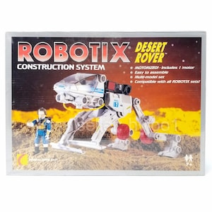 May include: A white and grey robotic toy with a clear cockpit, red accents, and a motor. The toy is labelled "ROBOTIX DESERT ROVER" and is part of a construction system. The toy is on a brown and orange surface.