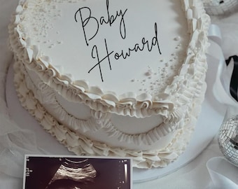 Könnte beinhalten: Ein weißer herzförmiger Kuchen mit den Worten "Baby Howard" in schwarzer Kursivschrift auf der Oberseite. Ein Schwarzweiß-Ultraschallbild befindet sich im Vordergrund.