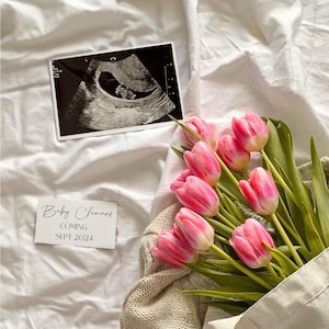 May include: A white card with the text "Baby Clemons Coming Sept 2024" on a white bedspread with a black and white ultrasound image and a bouquet of pink tulips in a white canvas bag.