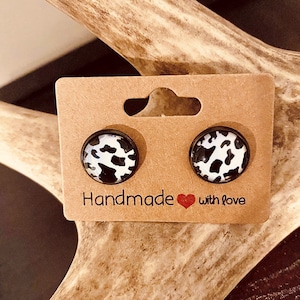 May include: A pair of round stud earrings with a black and white cow print design. The earrings are displayed on a brown card with the text "Handmade with love".