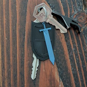 May include: A black 3D printed shield keychain with a blue plastic sword attached. The keychain is attached to a set of keys with a bottle opener.