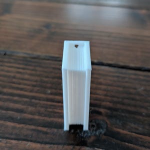 May include: A small, white, rectangular object with a vertical design. The object has a hole at the top and is standing upright on a wooden surface. The object appears to be 3D printed.