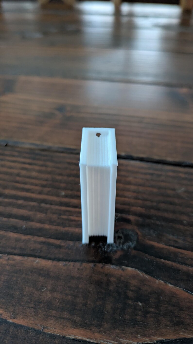 May include: A small, white, rectangular object with a vertical design. The object has a hole at the top and is standing upright on a wooden surface. The object appears to be 3D printed.