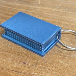 May include: A blue rectangular keychain resembling a miniature book. The keychain has a silver metal ring attached to the side. The item is made of a solid, possibly 3D-printed material.