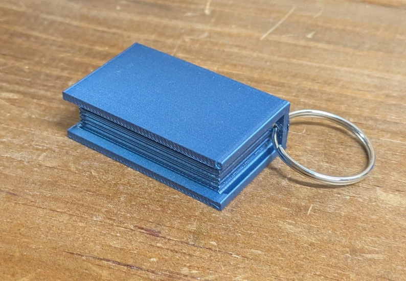 May include: A blue rectangular keychain resembling a miniature book. The keychain has a silver metal ring attached to the side. The item is made of a solid, possibly 3D-printed material.