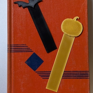 Pode incluir: Um livro laranja com um design de losangos e listras azuis, com um marcador de morcego preto e abóbora amarela. Os marcadores são colocados na capa do livro, sugerindo um tema de Halloween ou outono.