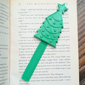 May include: Green Christmas tree-shaped bookmark made of plastic, resting on the pages of a book. The bookmark has a tree design with small, round details. The book is open on a wooden surface.
