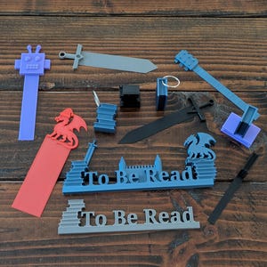 May include: A selection of 3D-printed bookmarks in various shapes and colours, including a robot, swords, a guitar, and dragons. Some bookmarks have the text "To Be Read". The items are arranged on a dark wood surface.